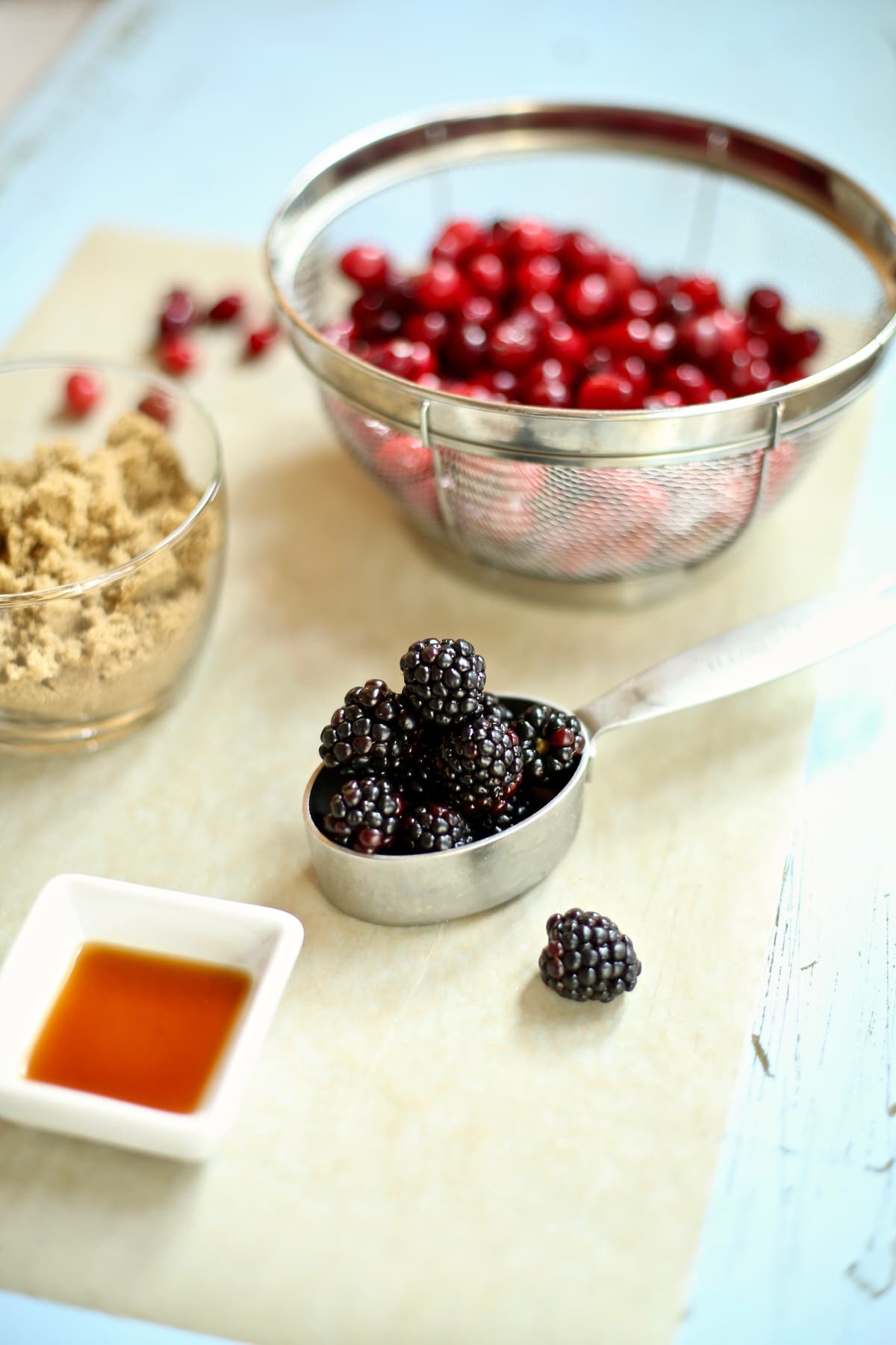 fresh blackberries in a silver measuring cup, with a glass cup of brown sugar next to it and a wire colander of cranberries behind it. 