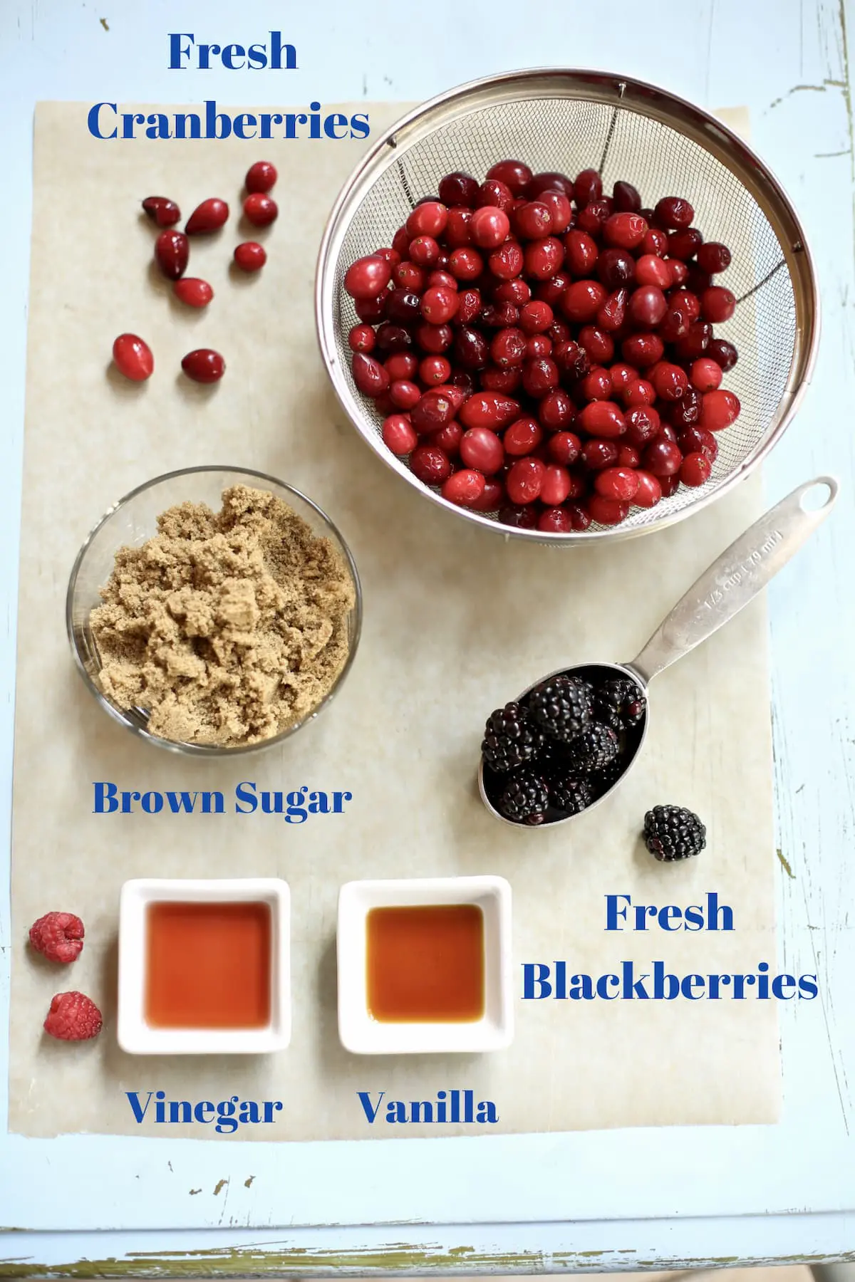 ingredients for cranberry sauce: fresh cranberries, brown sugar, raspberry vinegar, fresh blackberries on a blue table, with text.