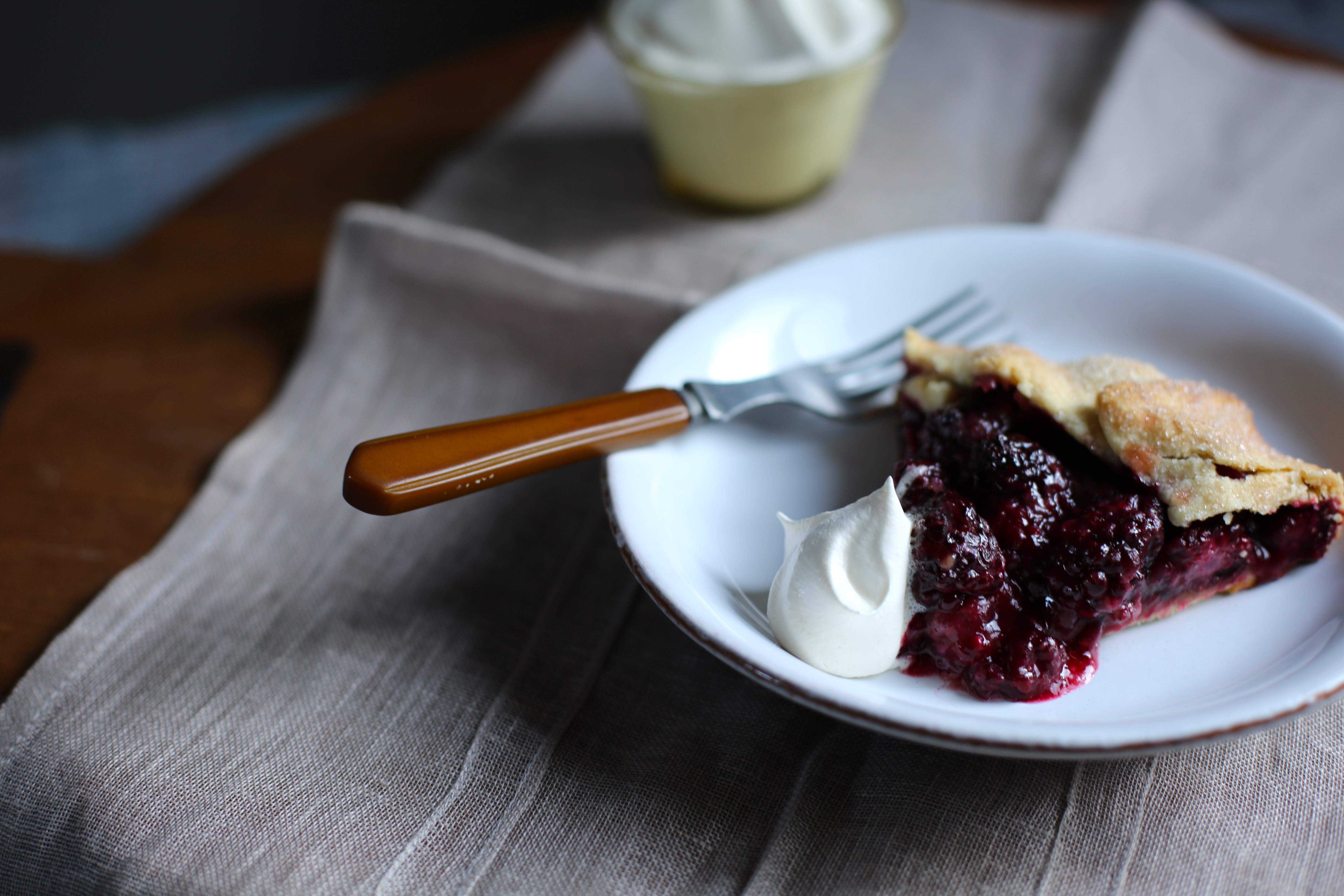 a slice of blackberry galette on a white plater and a brown fork with a dollop of whipped cream 