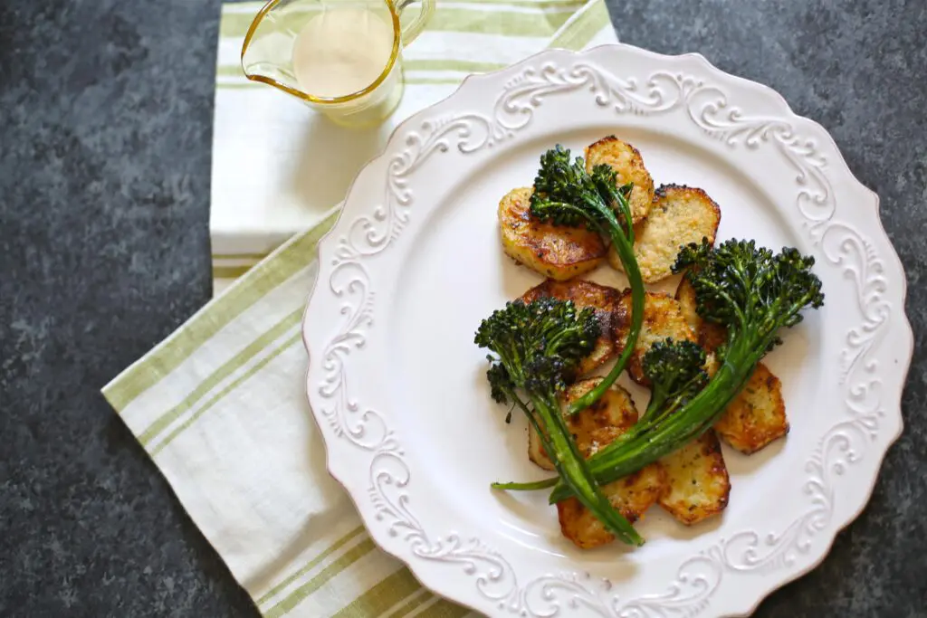 Potatoes and broccolini on a white platter with a sauce in a small yellow pitcher next to it.  On a gray table with a striped napkin. The image shows the dish without the sauce on top, that it can be served from the side. 