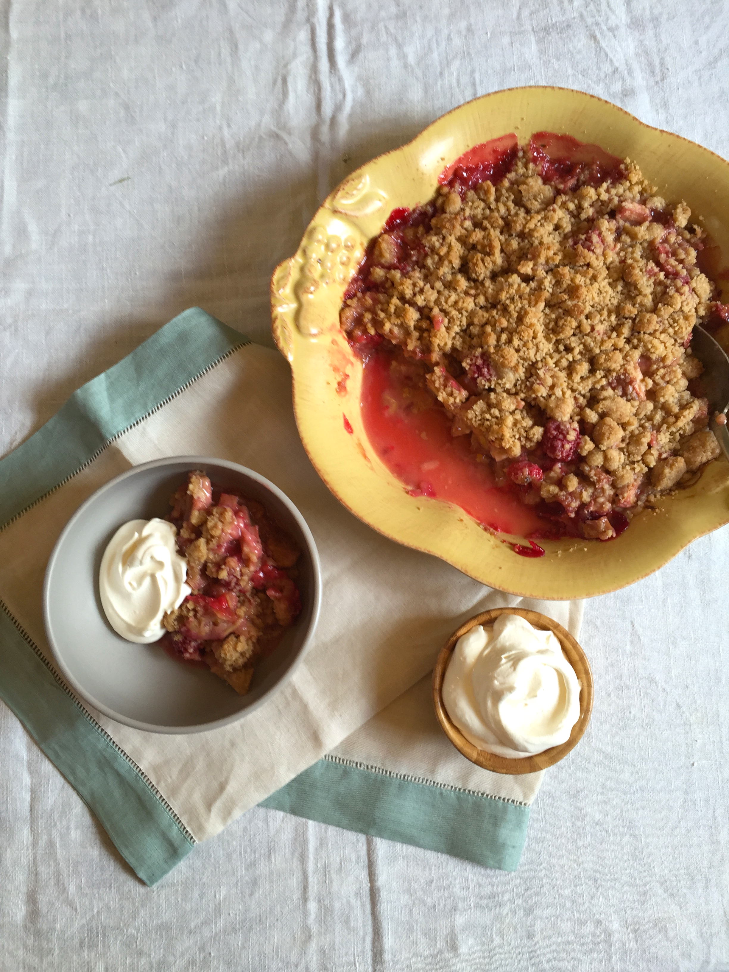 a yellow baking dish of Raspberry Rhubarb Crisp, and some in a bowl next to it with shipped cream on top. A small bowl of whipped cream sits beside it. 