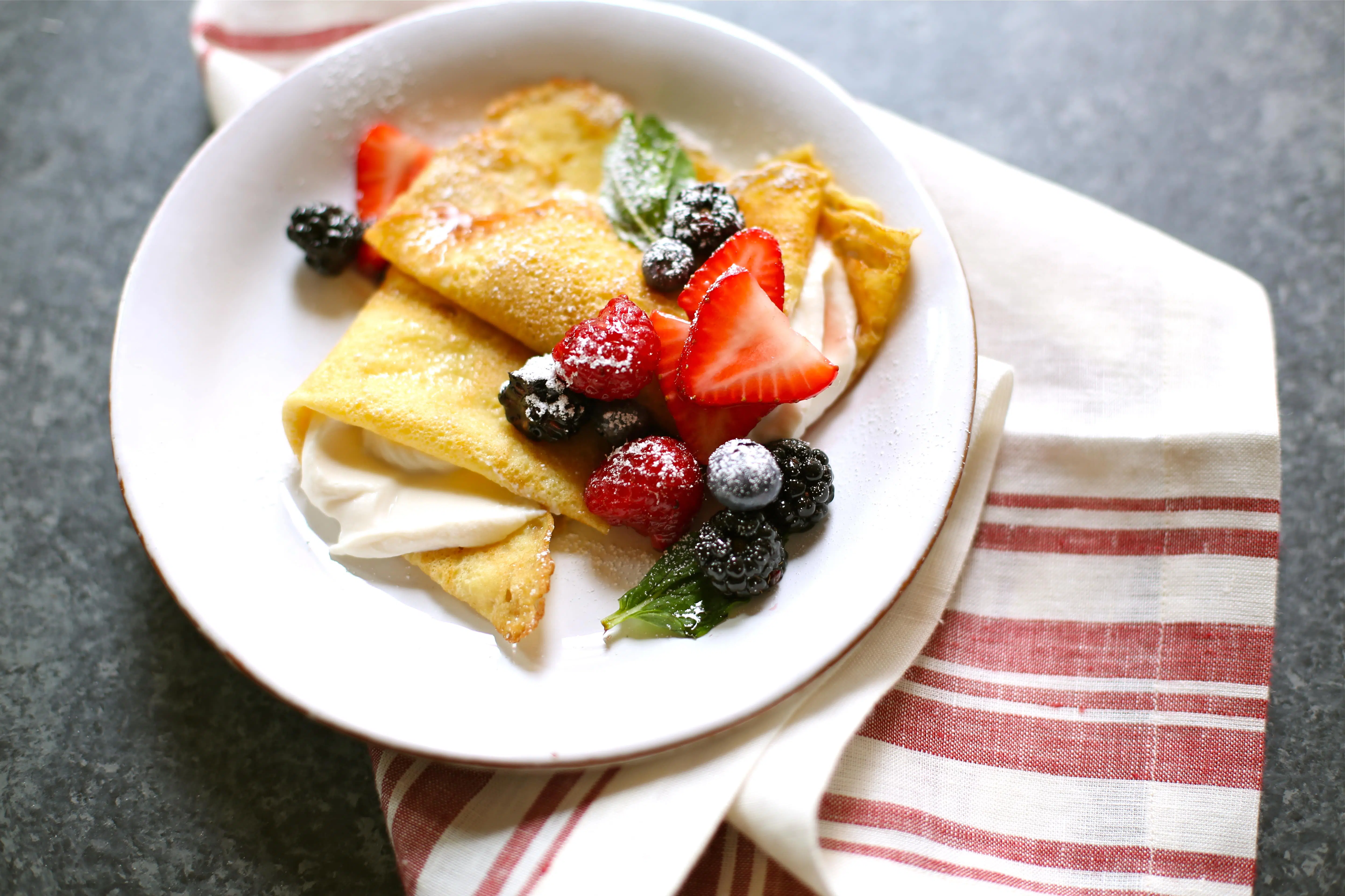 a plate of crepes with fresh fruit and cream on a black table with a striped red napkin.  