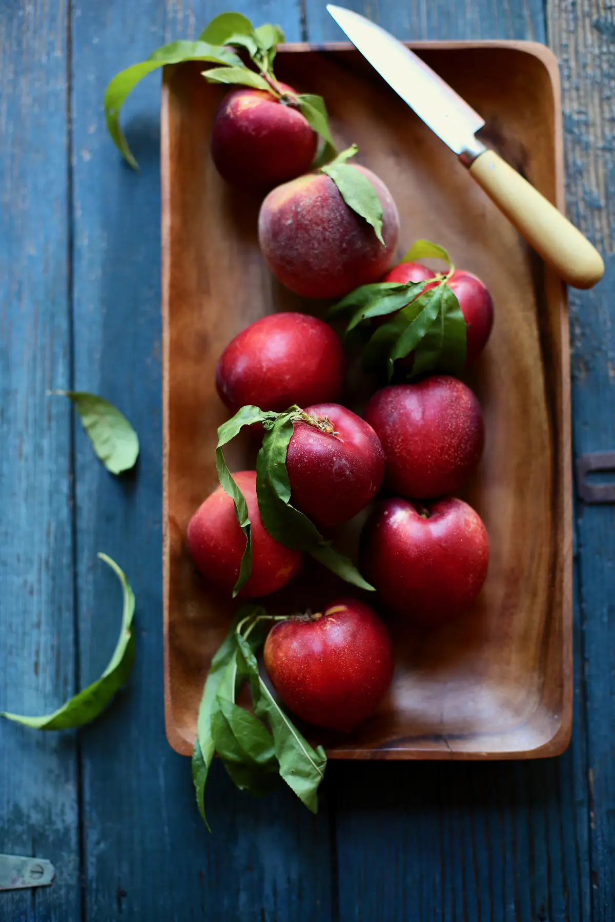 A wooden tray of nectarines on a blue table with a knife sitting next to it 