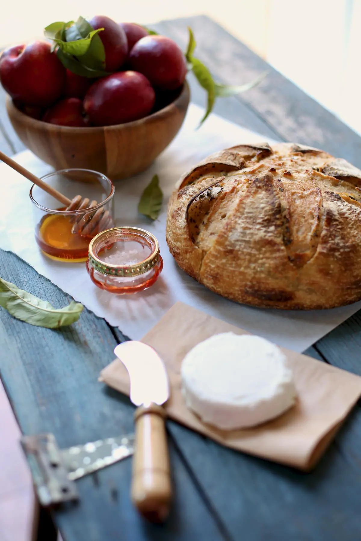 Fresh bread and goat cheese and honey on a table with a honey dipper and cheese spreading knife and small wooden bowl.