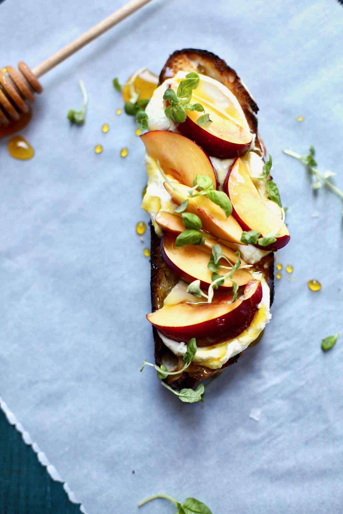 a big piece of toast with nectarine, goat cheese and honey on a parchment paper lined blue table