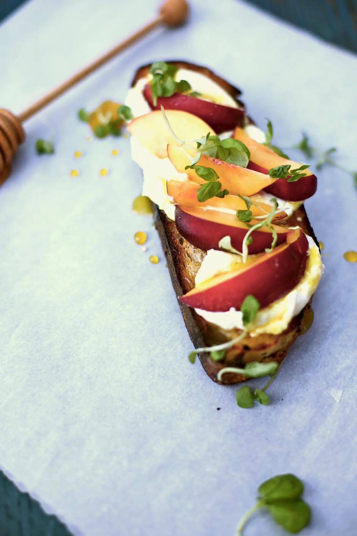 a big slice of toast with nectarines and honey and goat cheese and basil  on a white table with a honey dipper sitting nearby. 