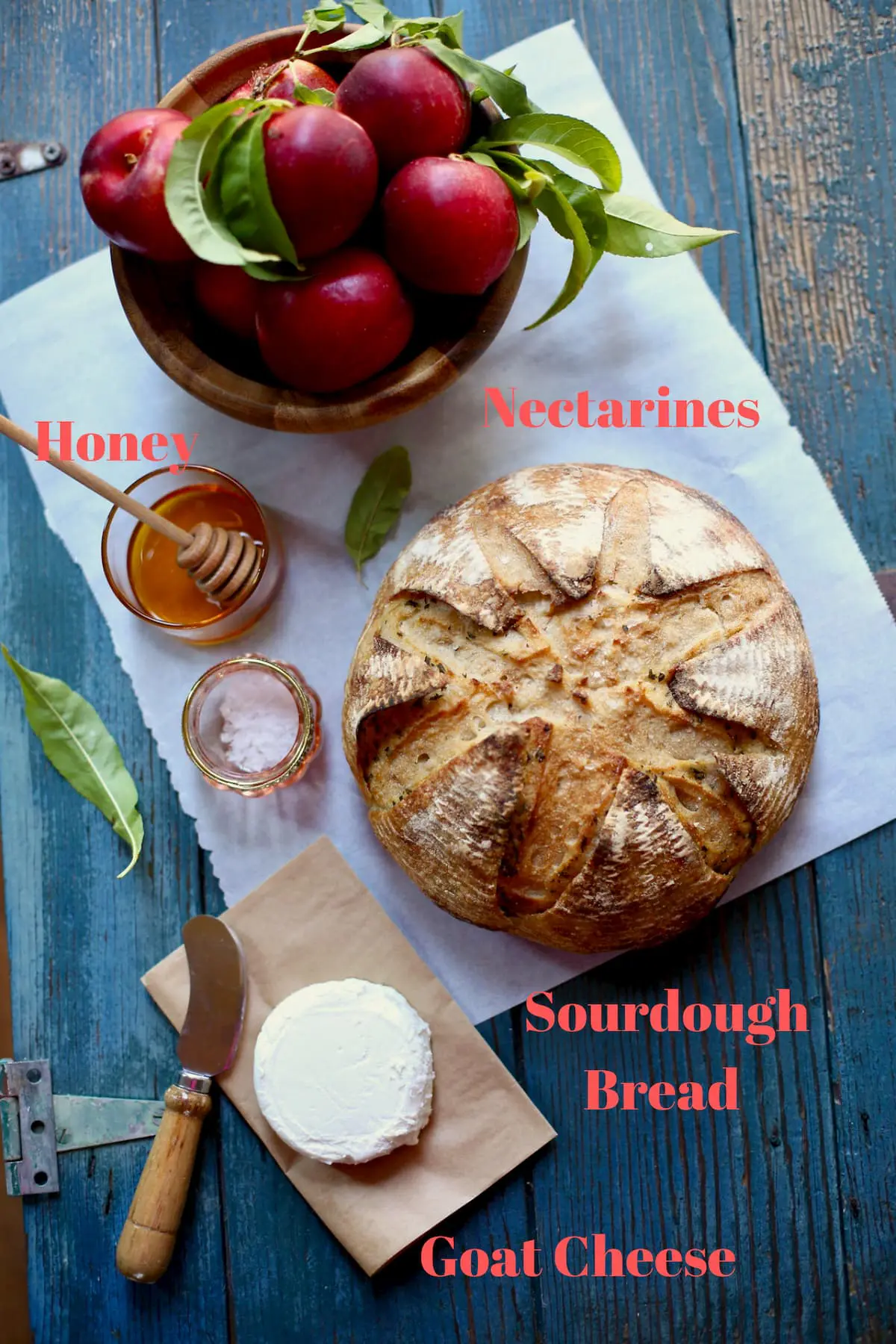 A round of bread, goat cheese, nectarines, honey, flaky sea salt and parchment paper sitting on a blue wooden table 