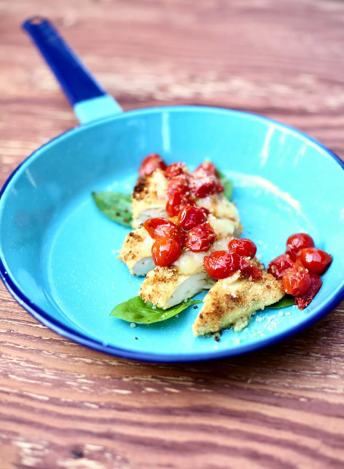 a side photo of chicken on a blue skillet with tomatoes on top and basil on the side.