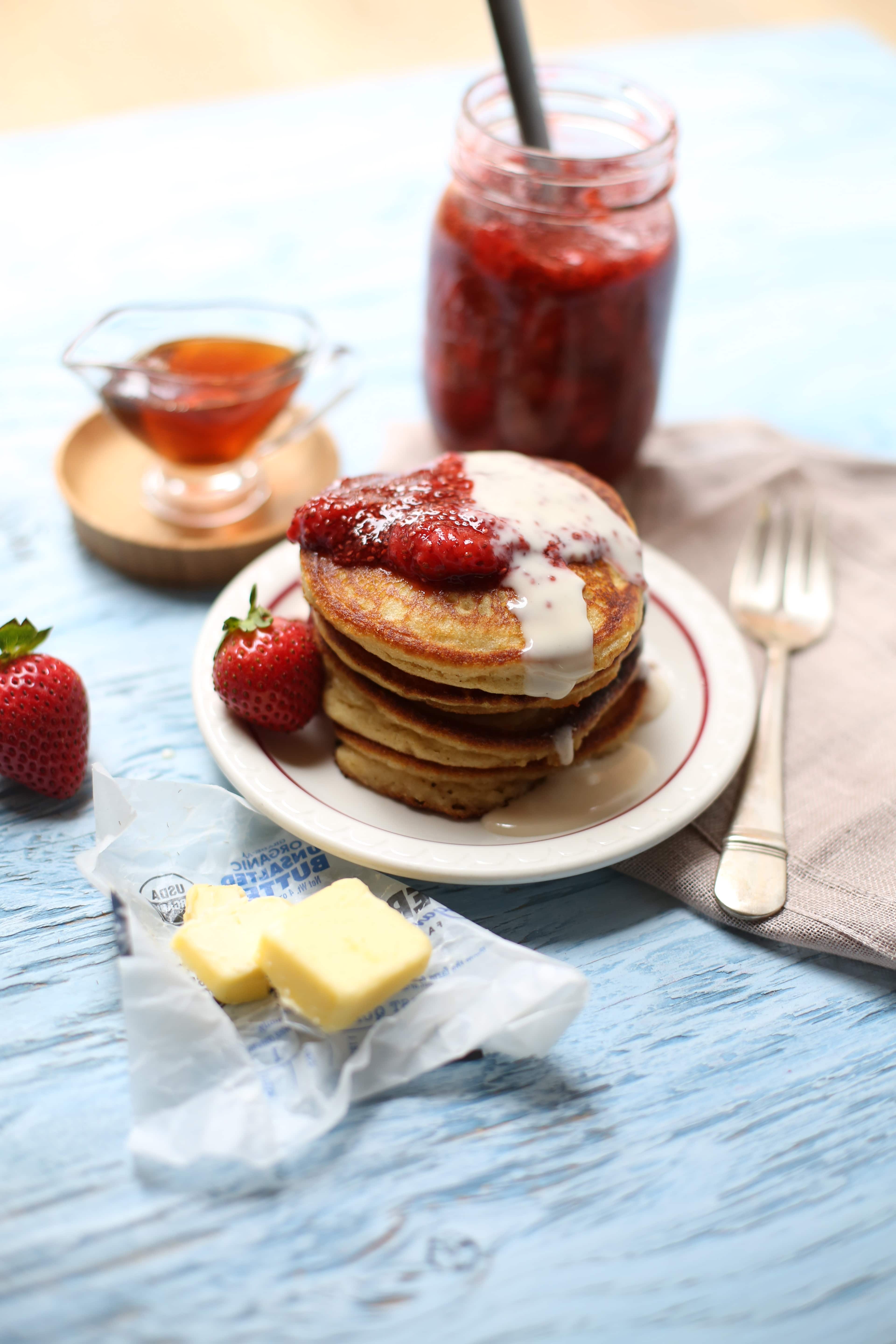 pancakes with strawberries on top and butter alongside with syrup and fork on a table. 