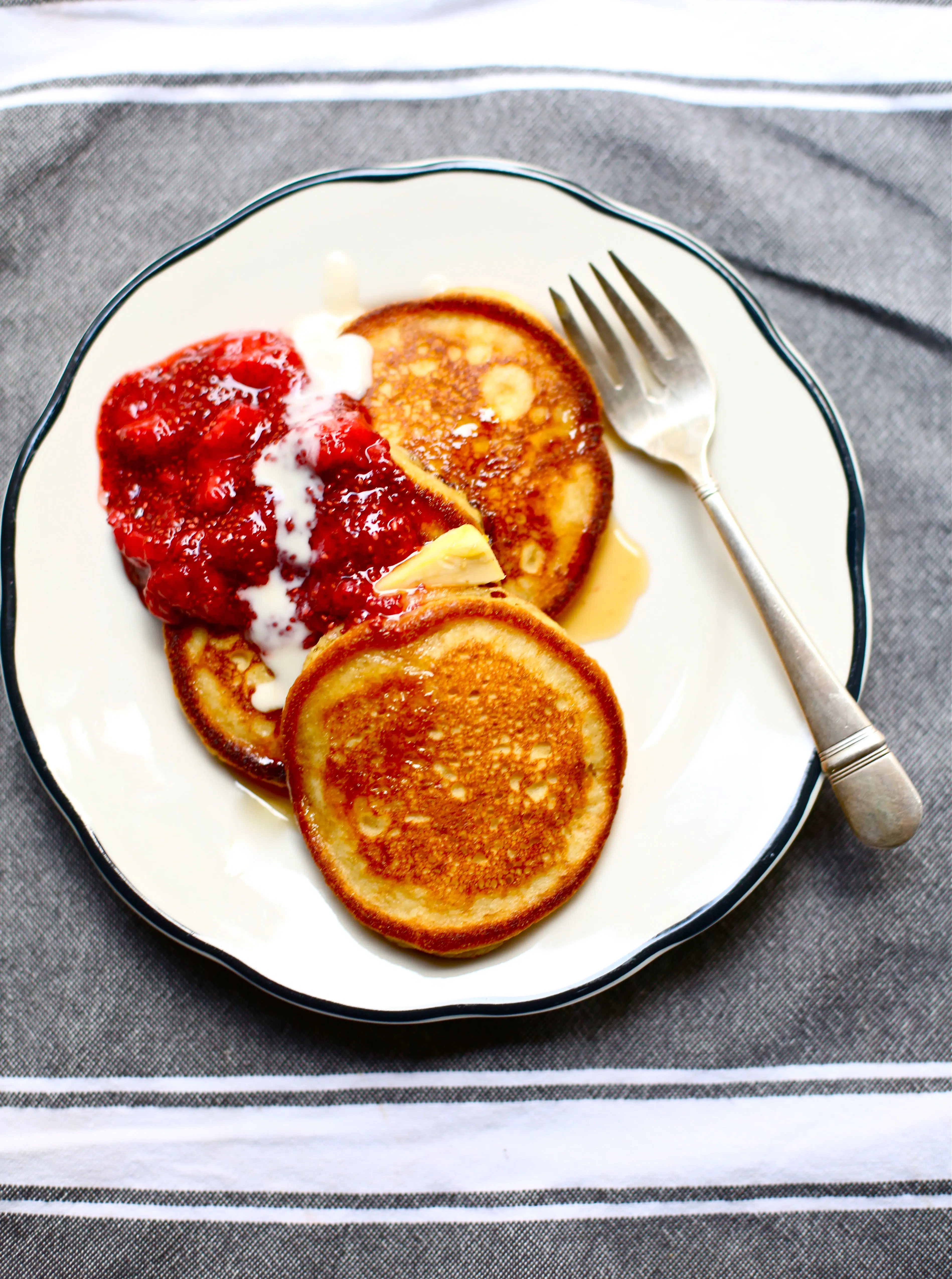 Cornmeal Pancakes with Strawberry Jam on a white plate with black rim and a fork sitting on gray and white background. 