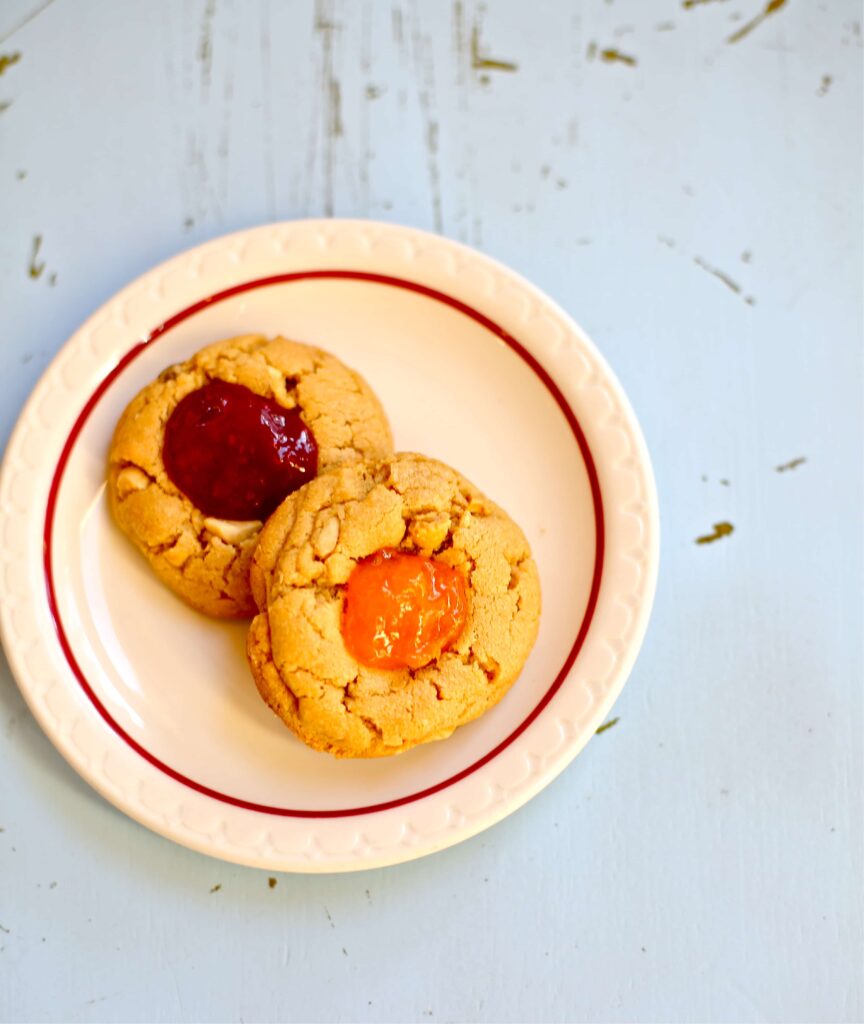 Two peanut butter and jam cookies on a white plate with red stripe, one cherry, one apricot 
