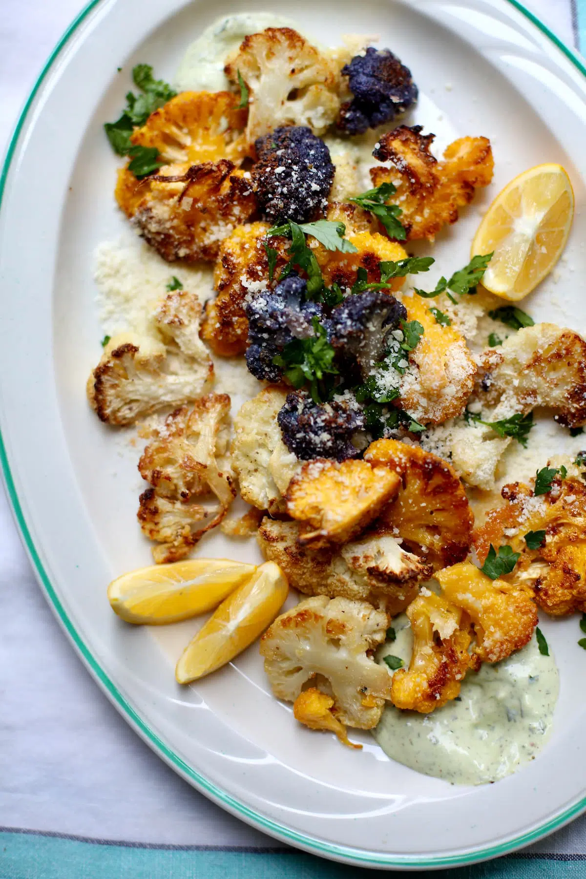 a white platter of roasted cauliflower with yogurt sauce under it and some lemon wedges on the side. 