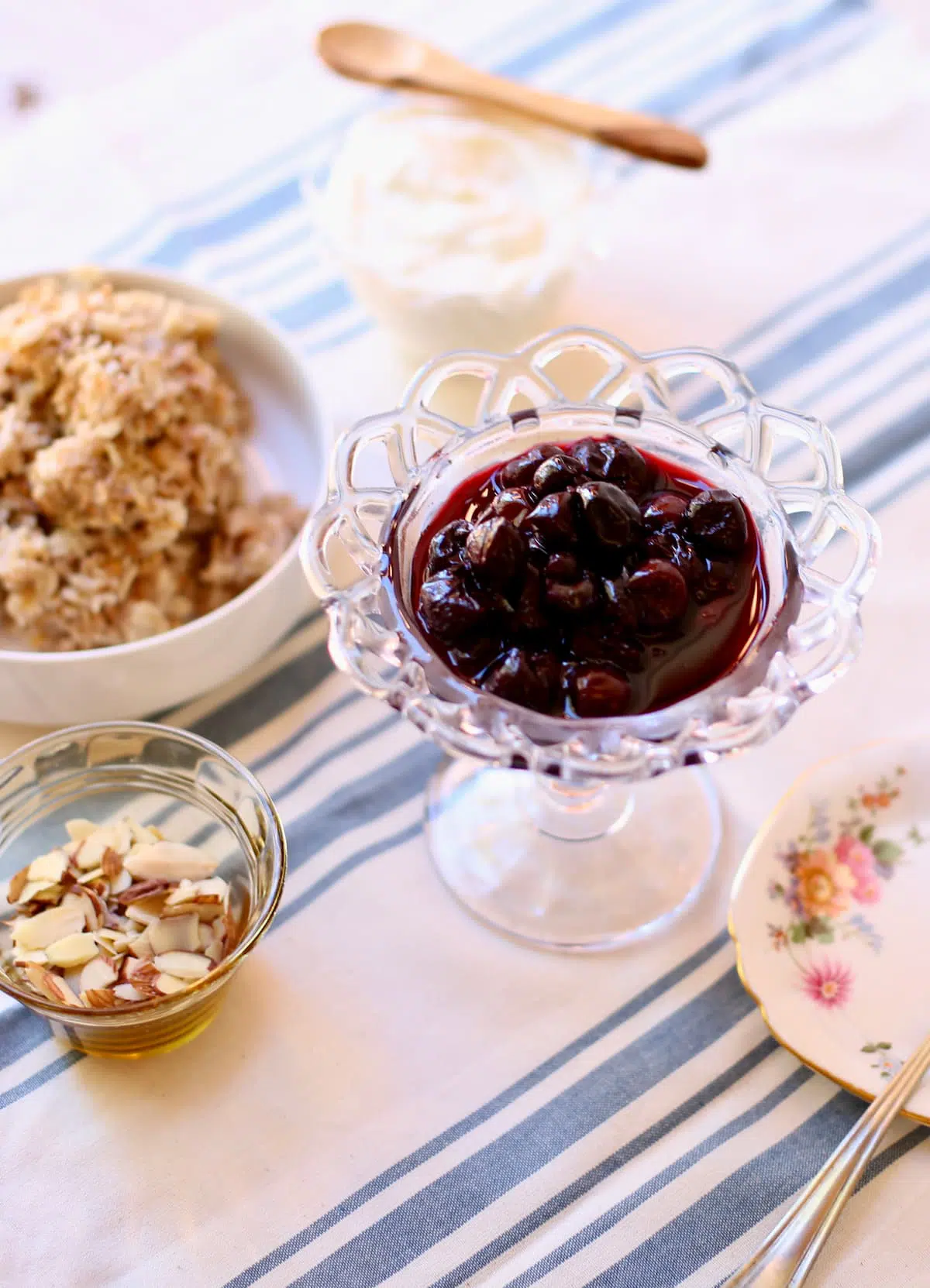 a table with a striped tablecloth, with a glass bowl of cherries, a bowl of oatmeal, some alomnds and a some yogurt.