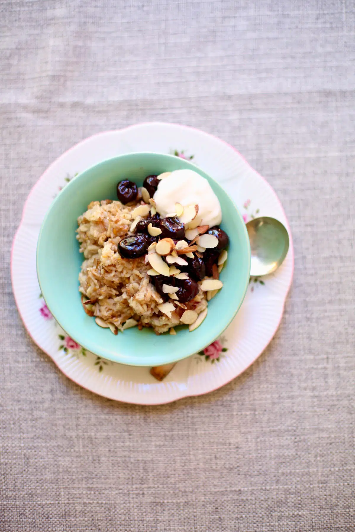a bowl of cherry oatmeal on a table with gray tablecloth, the saucer is green and there's a spoon alongside.