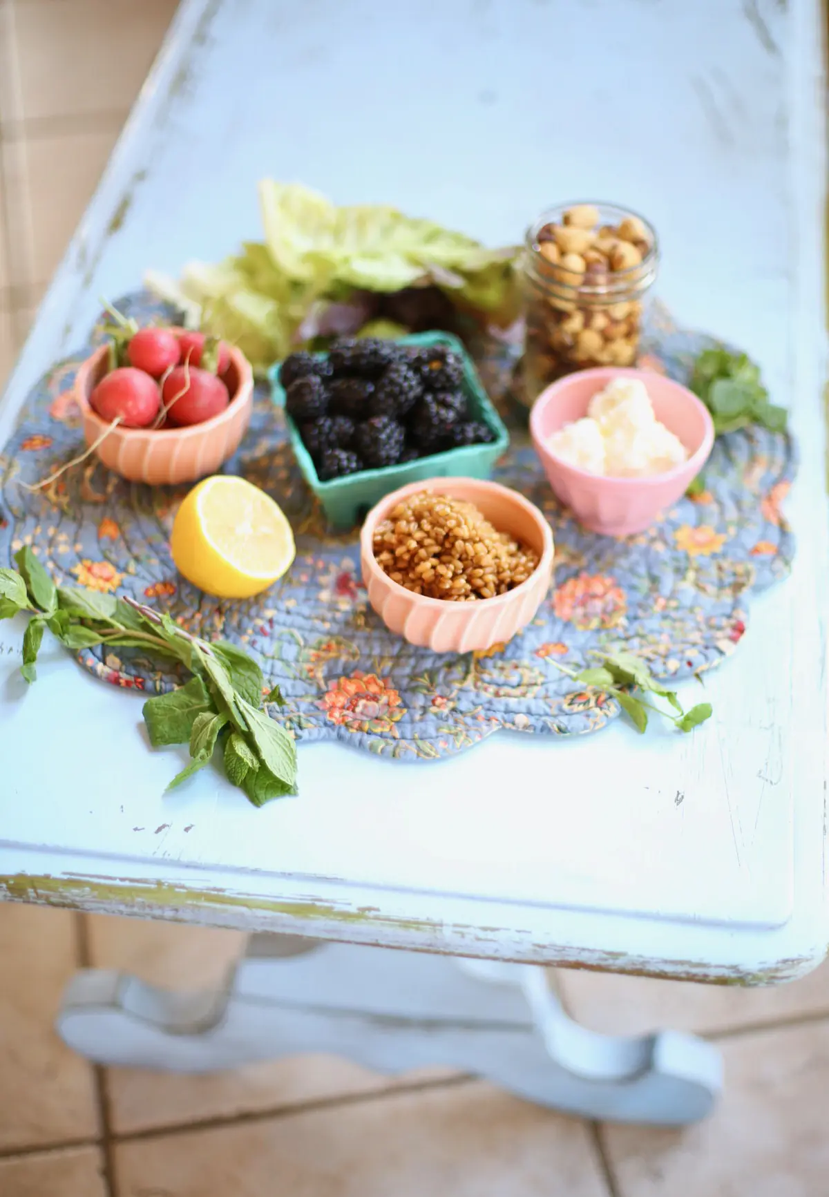 Ingredients for Blackberry Salad on a blue table: Grains, blackberries, hazelnuts, goat cheese, lemon, mint and radish in small bowls.