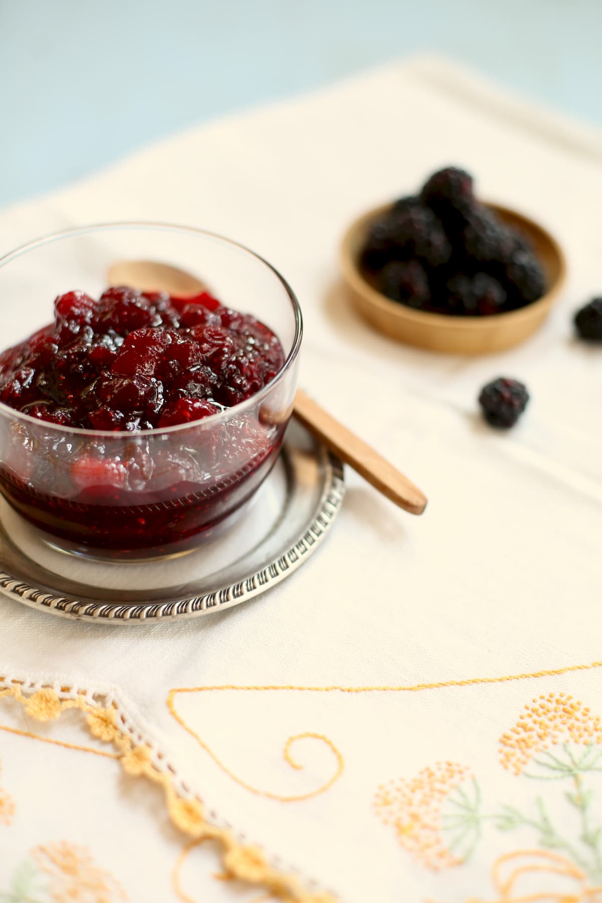 a glass bowl of cranberry sauce with blackberries behind it on a tablecloth with yellow trim.
