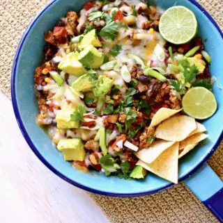 a blue skillet with cooked taco meat with melted cheese, chips, lime and avocado.