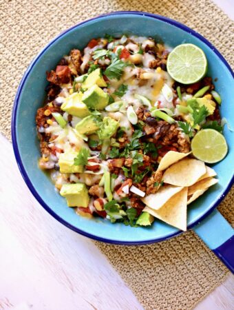 a blue skillet with cooked taco meat with melted cheese, chips, lime and avocado.