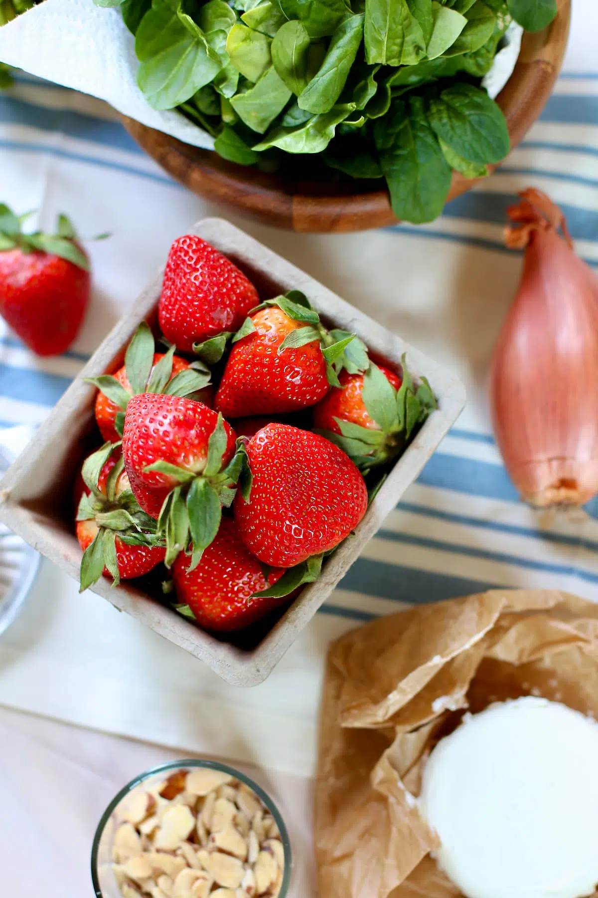 a basket of strawberries and a shallot and some spinach and other salad ingredients on a striped tablecloth.