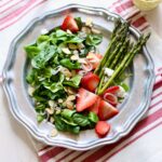 a photo of a strawberry goat cheese salad with asparagus on it, on a silver plate sitting on a striped tablecloth.