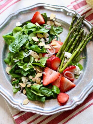 a photo of a strawberry goat cheese salad with asparagus on it, on a silver plate sitting on a striped tablecloth.