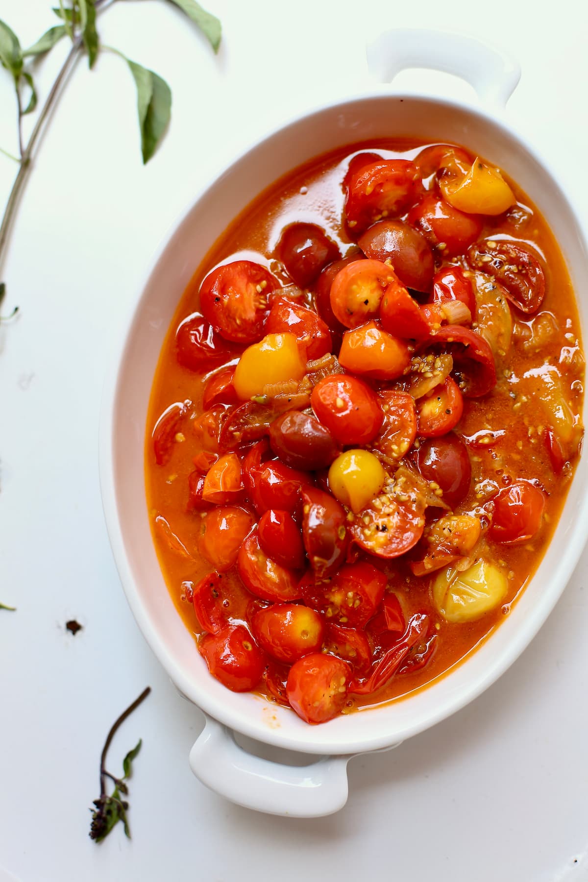 a white serving dish of cooked cherry tomatoes for a sauce on a white table.