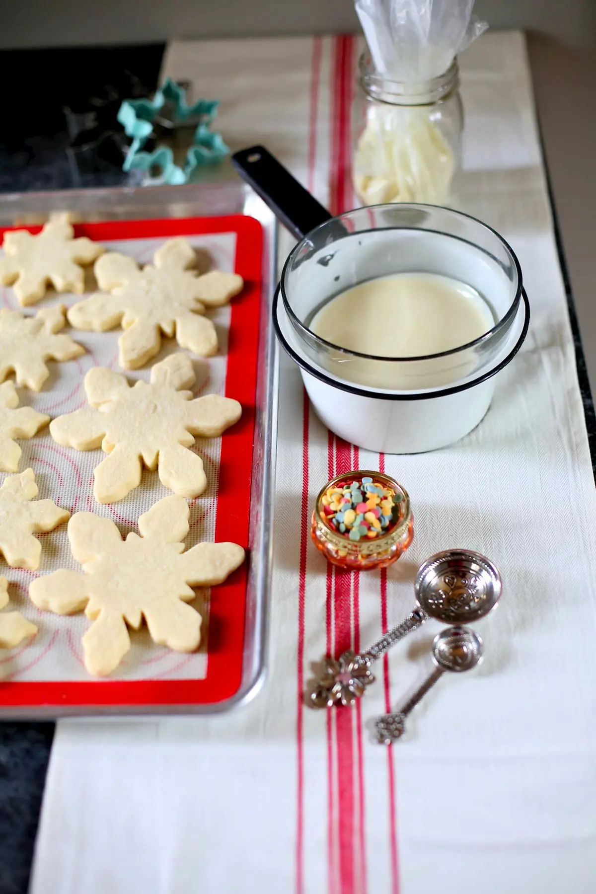 plain unfrosted snowflake cookies and melted white chocolate and sprinkles and measuring spoons on a table