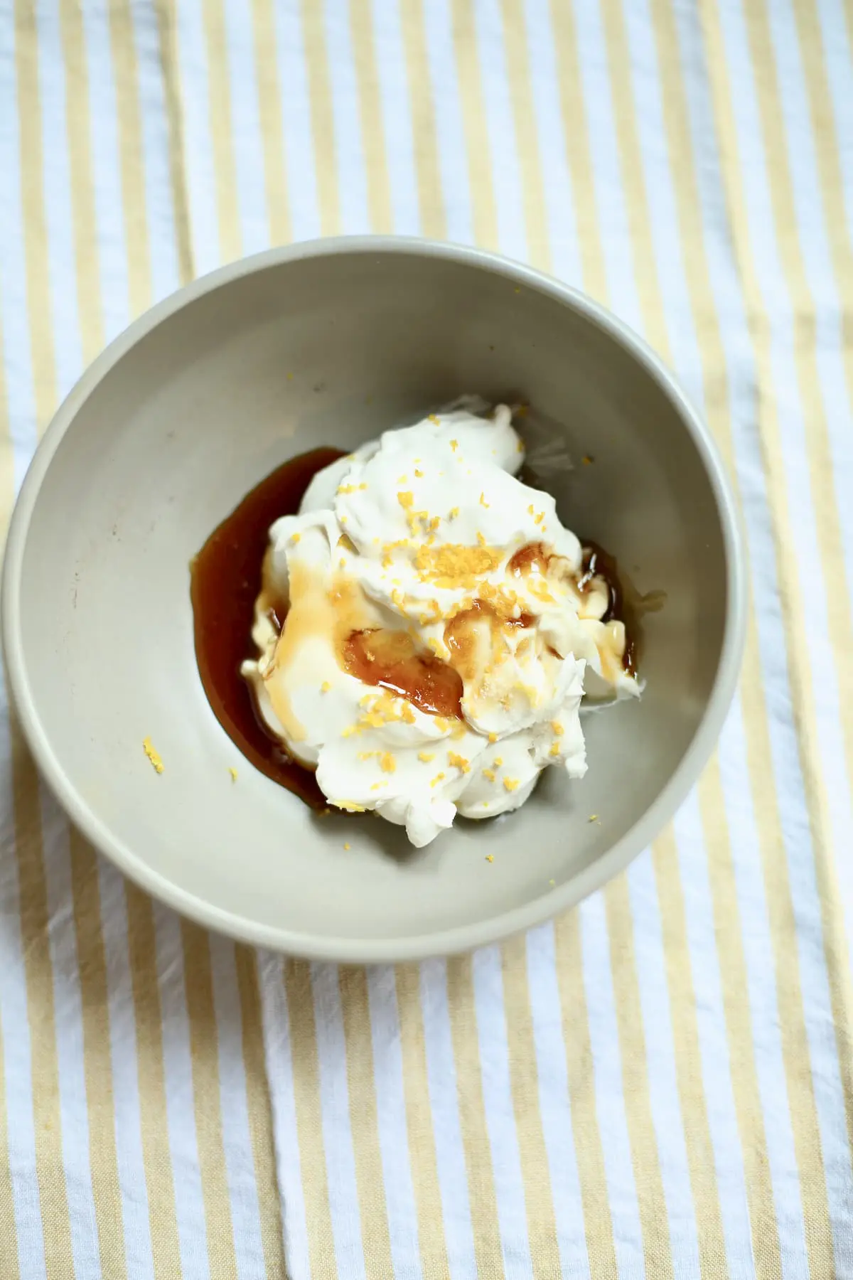 A small gray bowl of whipped goat cheese with lemon zest and honey on top on a yello striped tablecloth