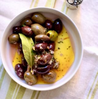 a bowl of olives with bay leaf and olive oil in a white bowl on a table