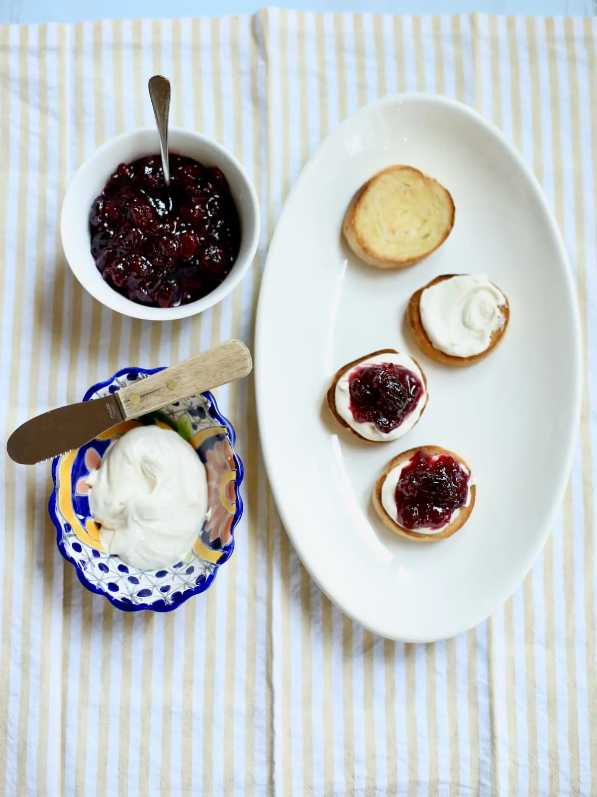 a white platter of partially made appetizer toasts and a small bowl of cheese and and a bowl of cranberries