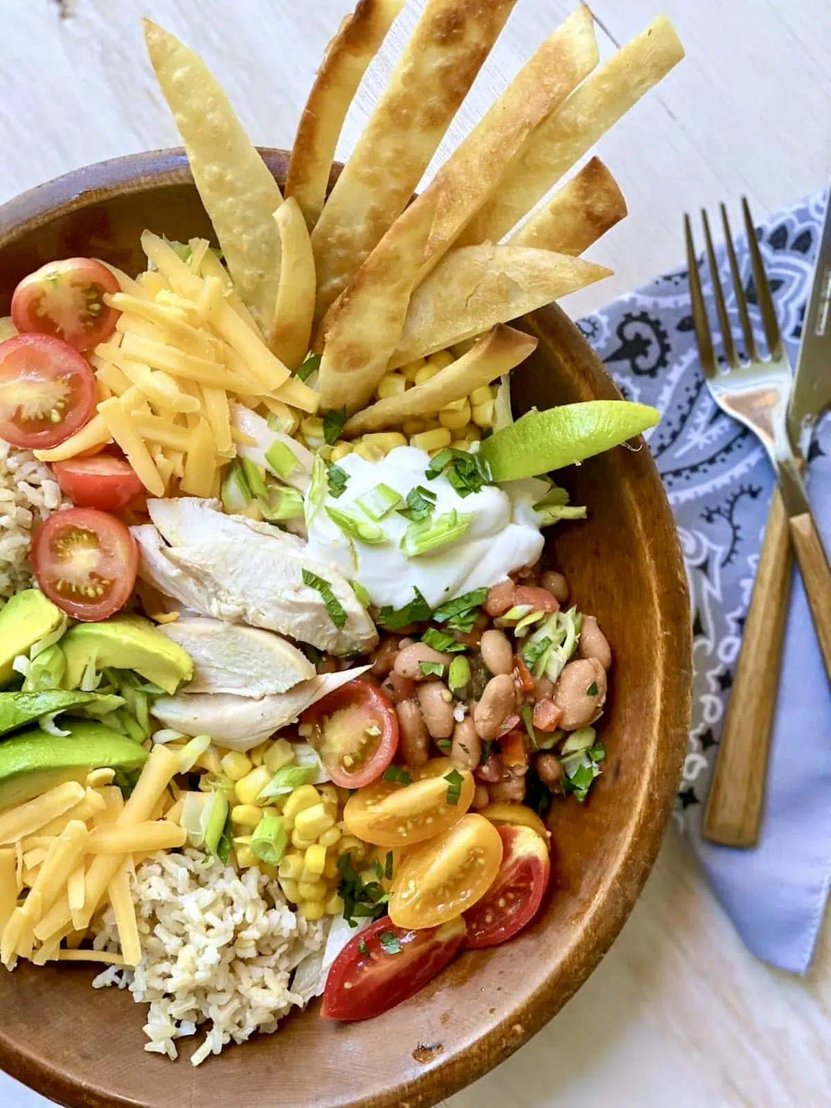 Closeup shot of a burrito bowl, tortillias, sour cream lime tomato cheese rice corn lettuce and a knife and fork