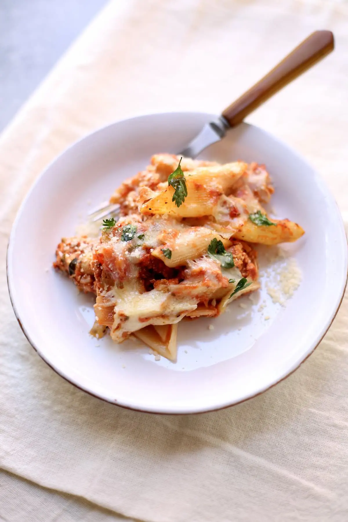 a white plate of baked pasta with a fork, sitting on a white tablecloth.