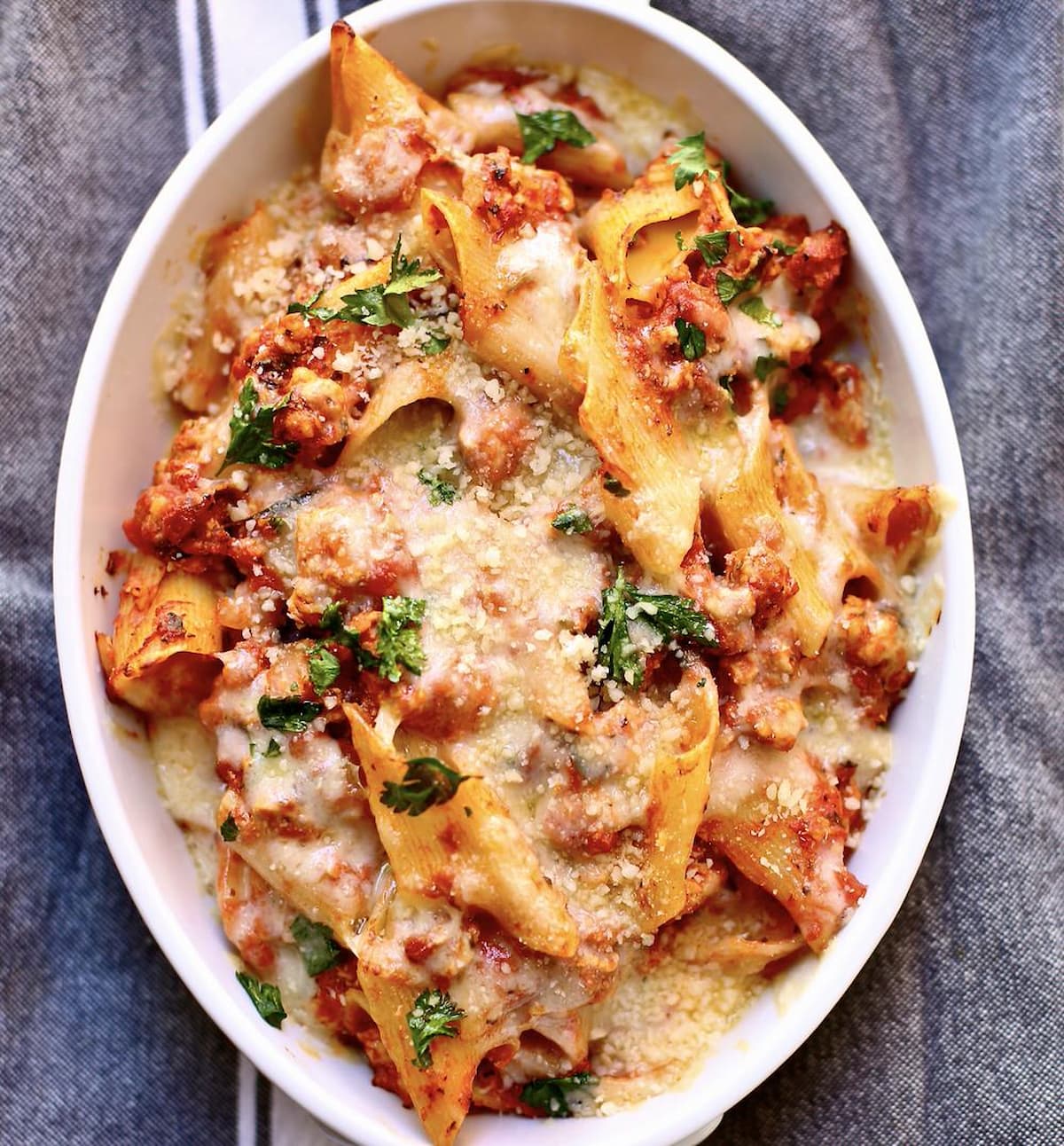 baked tomato and sausage pasta in a white casserole dish with melted cheese on top on a gray tablecloth 