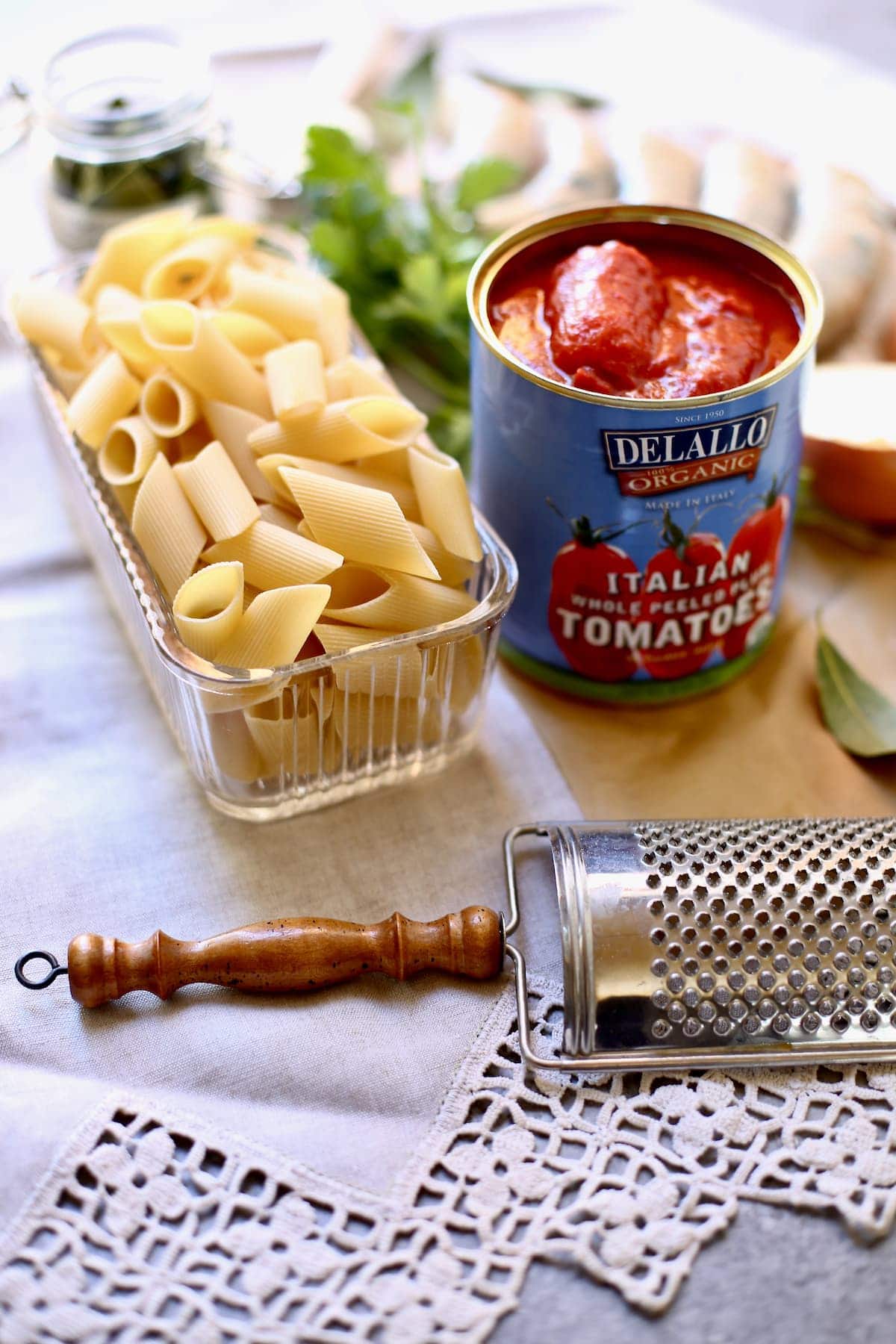 a can of tomatoes, pasta and a cheese grater on a lace tablecloth and gray table 