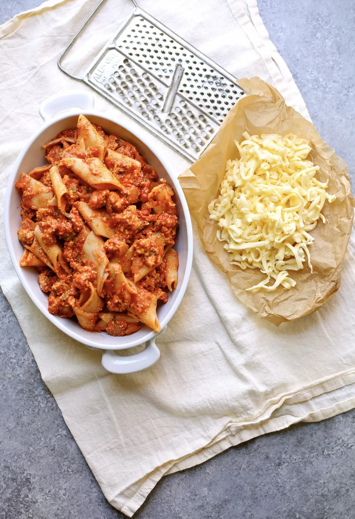 a casserole dish of pasta ready for the oven and some parchment paper with grated cheese next to it, ready to be added to the top and then put in the oven.