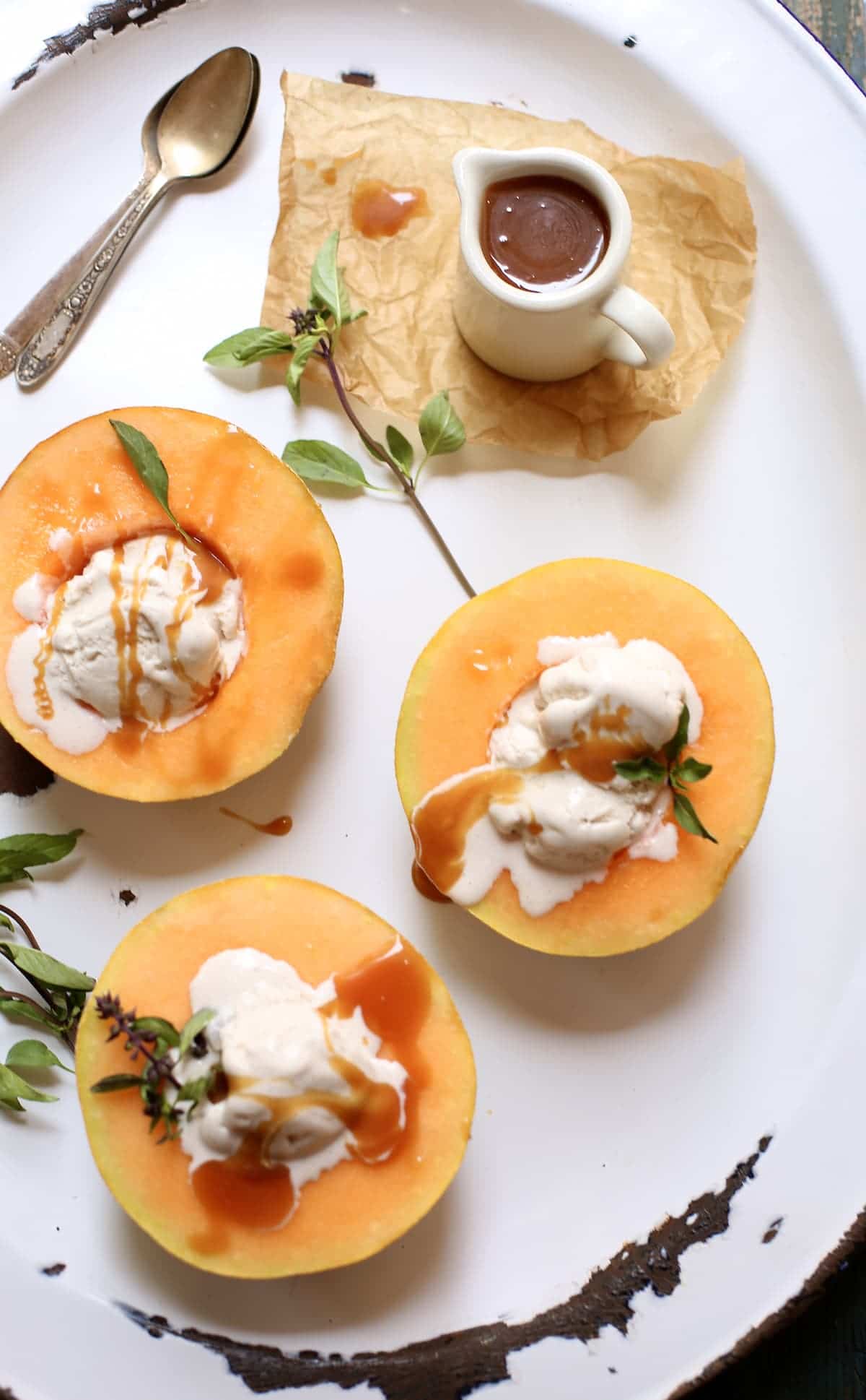 three orange melon halves with ice cream in the center and caramel sauce and a pitcher of caramel sauce and also spoons, sitting on a white platter.