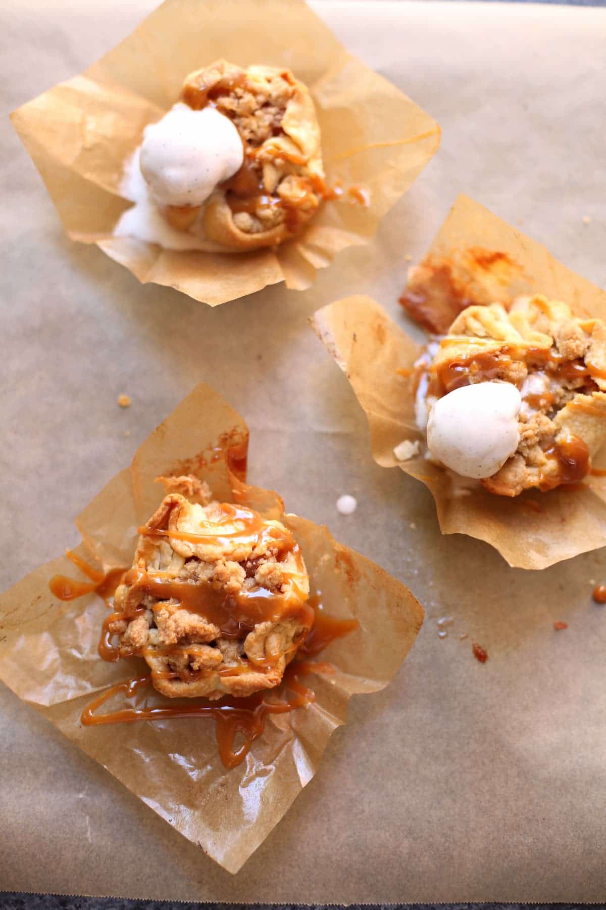 three small pies, two with ice cream on and all with caramel sauce on parchment paper.