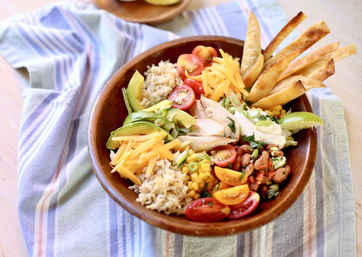 a wooden bowl of food: chicken, rice, tortillias, cheese, beans, salsa and avocado and tomato, on a blue tablecloth