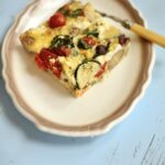 a piece of frittata on a white plate with beige trim with a fork on a blue table.