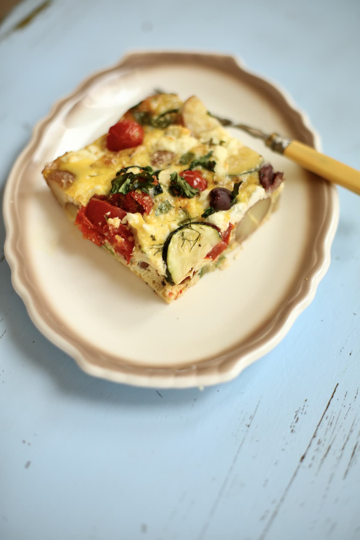 One slice of zucchini potato frittata on a white plate with beige trim, with a fork, sitting on a blue table.