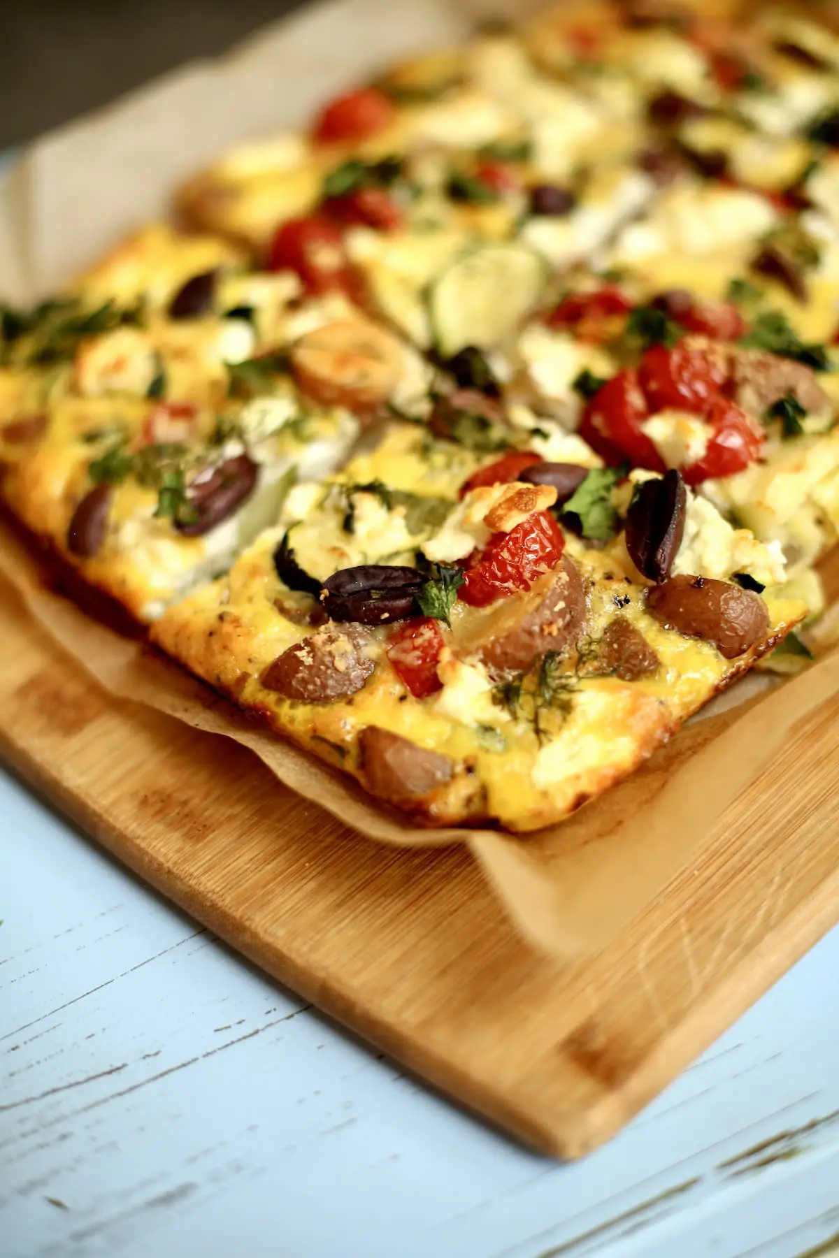 A wooden cutting board with a vegetable frittata sitting on it cut into slices.