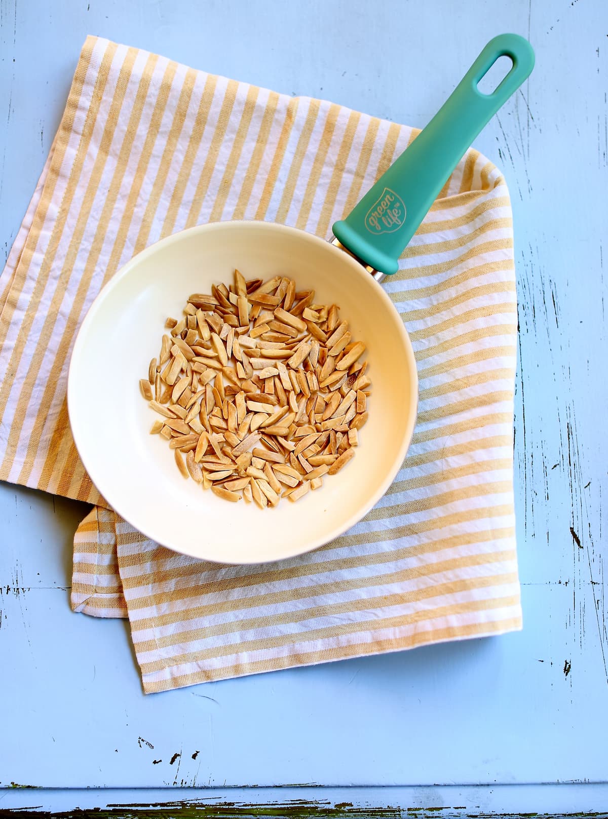 a white skillet with toasted slivered almonds inside on a striped cloth and blue table