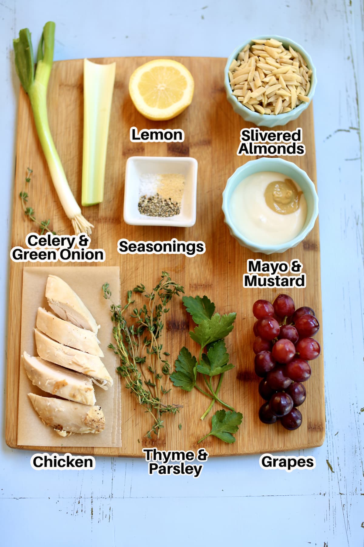 a cutting board of ingredients for chicken salad, rotisserie chicken, fresh herbs, grapes, mayo/mustard, seasonings, celery, lemon, slivered almonds.