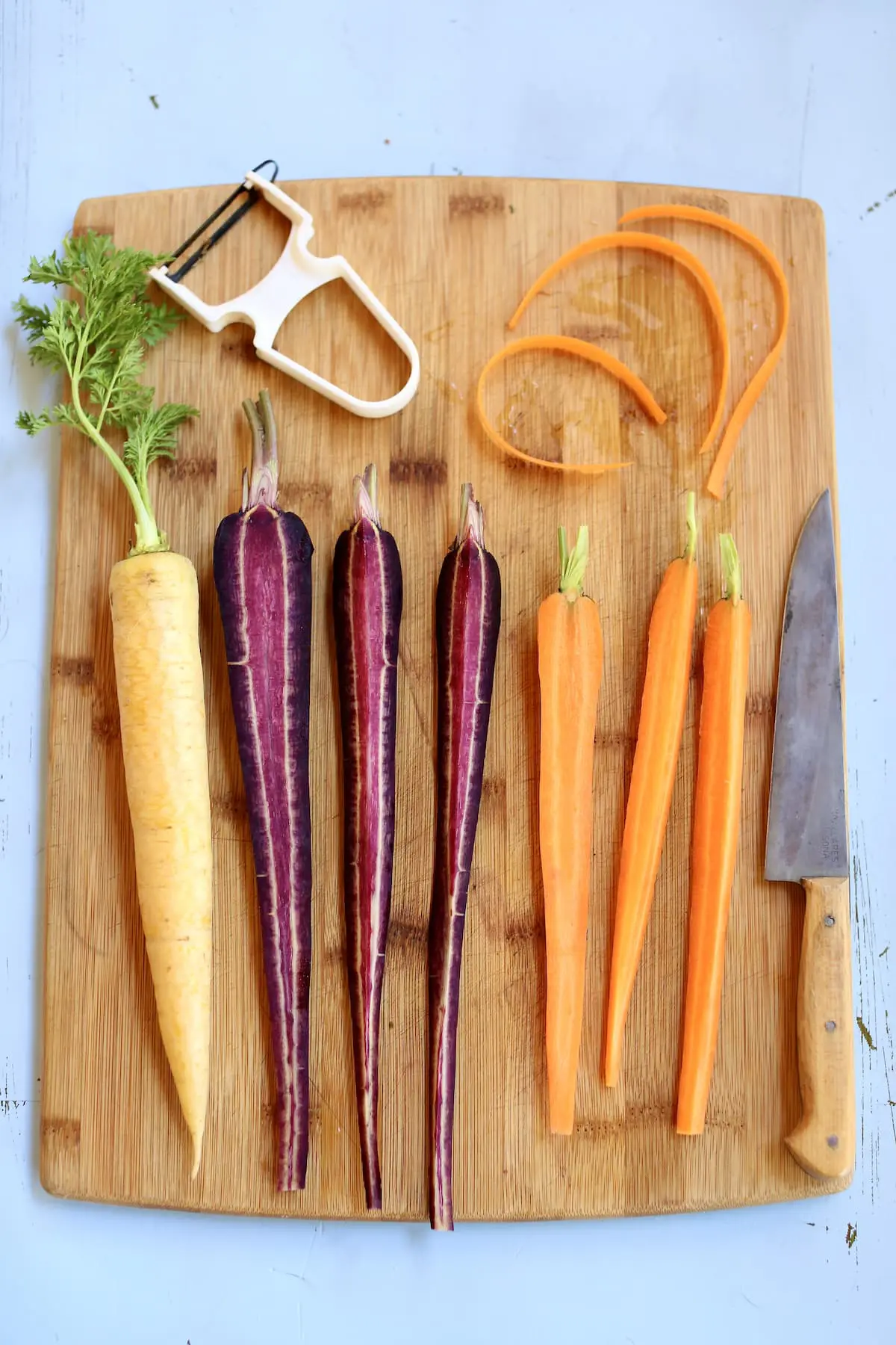 a cutting board with 7 carrots, yellow purple and orange, with a vegetable peeler and a knife.