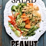 a white platter with peanut noodle salad on a blue table. black and white text are overlaid on the photo