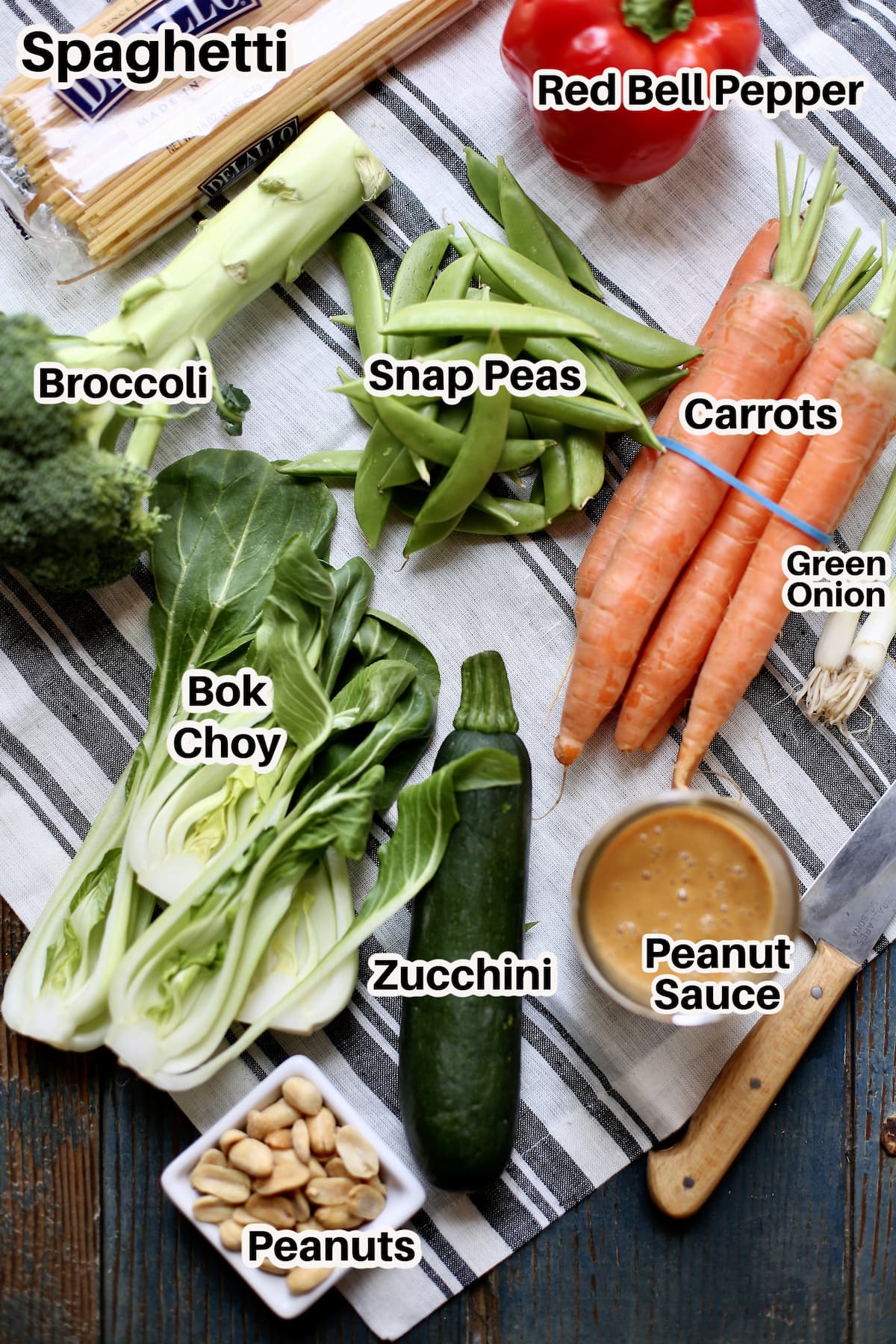 a table with striped cloth underneath with a text overlay saying the ingredients: snap peas, carrots, bok choy, zucchini, broccoli, peanut sauce green onion.