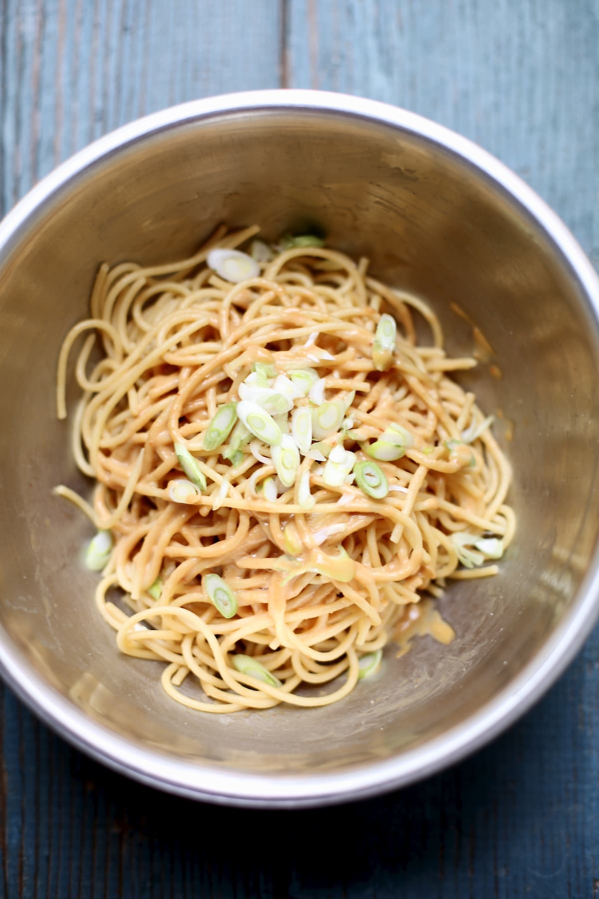 a silver bowl with pasta noodles and green onions on top on a blue wooden table. 