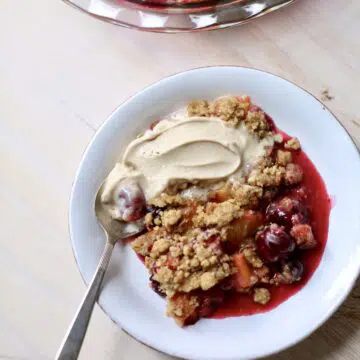 a white dish of summer fruit crisp with ice cream and a spoon on a table with a larger dish of it in the back.