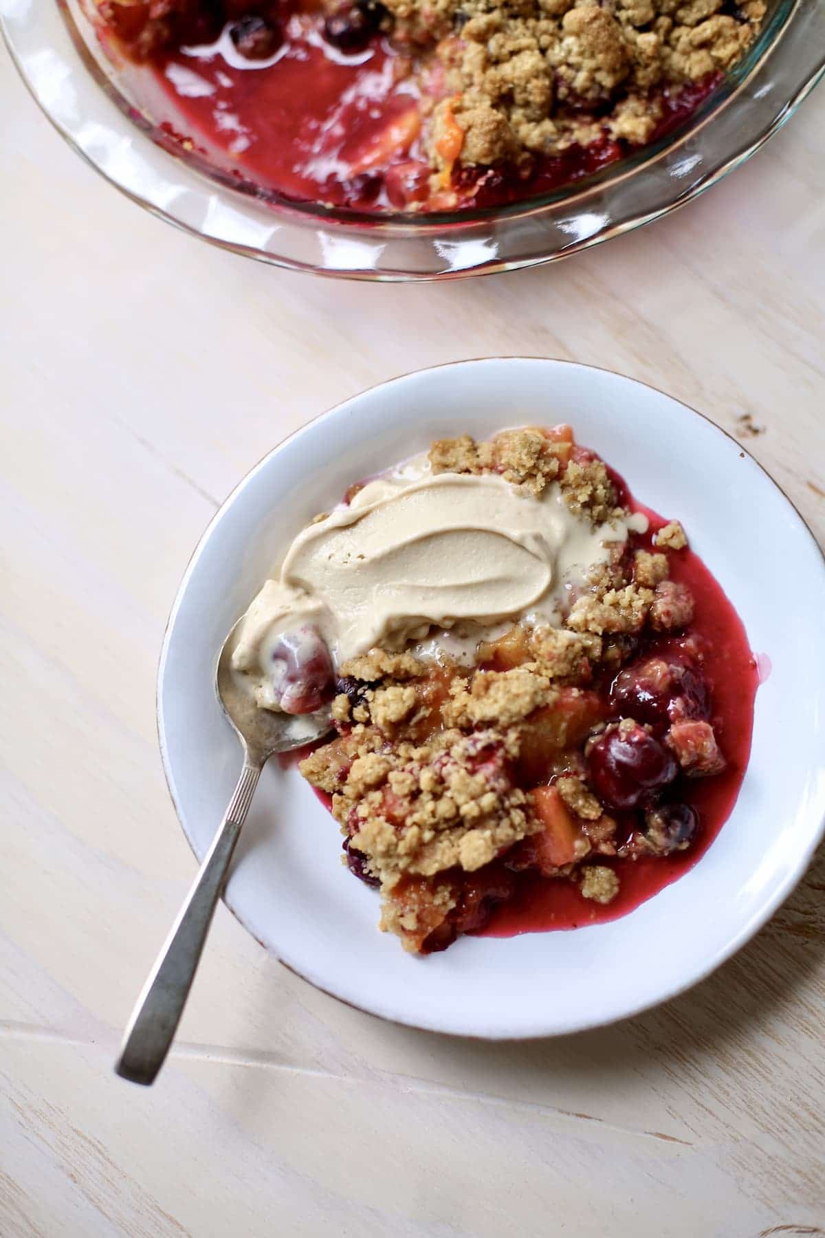 a white dish of summer fruit crisp with ice cream and a spoon on a table with a larger dish of it in the back.