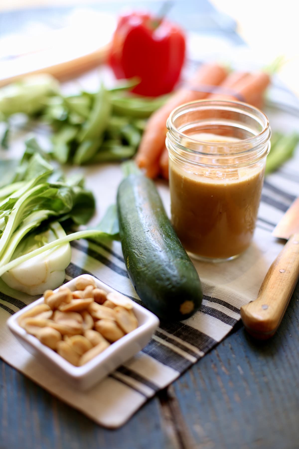 a table with a striped tablecloth and a canning jar of peanut sauce, surrounded by fresh vegetatables and a small bowl of peanuts